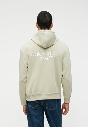 Light gray hoodie with a drawstring hood, featuring a white "Calvin Klein JEANS" logo on the back. Soft fabric and ribbed cuffs. Jeans visible below.