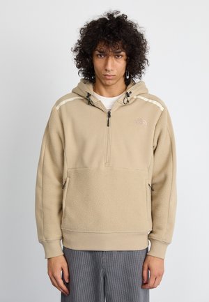 Sudadera polar beige con media cremallera, capucha ajustable y bolsillos laterales. Presenta una sección inferior texturizada y un sutil detalle del logo en el pecho.