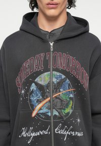 Felpa nera con zip che presenta una stampa grafica della Terra e di un angelo, con il testo "SOMEDAY TOMORROW" e accenti di stelle e di una cometa.