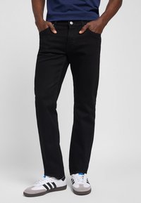 Homme portant un jean noir droit et des baskets blanches avec des rayures noires, mains dans les poches, sur un fond clair uni.