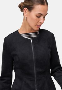 Giacca in suede nera con collo alto e zip anteriore. Presenta maniche lunghe e una vestibilità sartoriale. Sotto, è visibile una maglietta a righe.