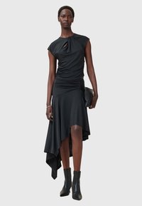 Schwarzes asymmetrisches Kleid mit kurzen Ärmeln, gerafftem Material und einer Twist-Detail am Hals; kombiniert mit schwarzen Knöchelstiefeln.