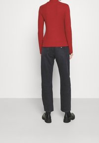 Person in einem roten Langarmshirt, schwarzen Jeans und schwarzen Stiefeletten, der mit dem Rücken zu einer einfachen hellen Hintergrund steht.