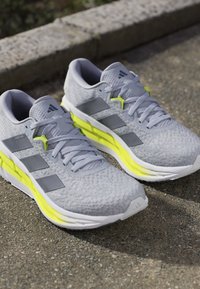 Zapatillas deportivas de color gris claro con una parte superior texturizada, que cuentan con tres franjas plateadas, detalles en amarillo y una suela acolchada. Diseñadas para la comodidad.