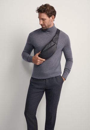 Mężczyzna w szarym golfie i ciemnych spodniach trzyma czarną skórzaną torebkę na ramię z zamkiem błyskawicznym na klatce piersiowej na jednolitym tle.