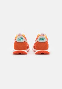 Ett par orange Nike-sneakers sedd bakifrån, med gröna "nike"-texter på vita hältabbar och texturerade sulor.
