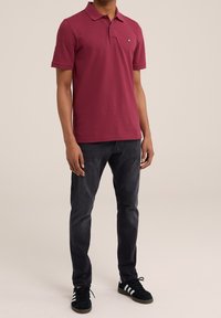 Bordový polo tričko s krátkým rukávem, límečkem a vyšitým logem, spárované s tmavými džíny a černými teniskami s bílými pruhy.