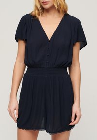 Marineblå ærmeløs romper med V-hals, knapdetaljer, smocket liv og bølget kant. Fremstillet af let, struktureret stof.