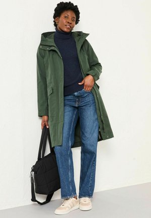 Grüne wasserdichte Jacke mit Kapuze, marineblauer Strickpullover, Jeans in Blau und hellfarbige Sneakers. Schwarze Schultertasche mit Reißverschluss.