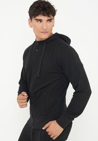 Felpa zip nera con cappuccio e lacci, maniche lunghe, tasche a marsupio e design aderente. Tessuto liscio con sottili dettagli di texture.