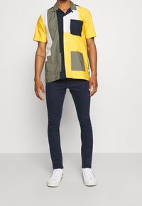 Camisa de manga corta de múltiples colores con bloques amarillos, verdes, blancos y azul marino, con un bolsillo. Combinada con jeans oscuros ajustados y zapatillas blancas.