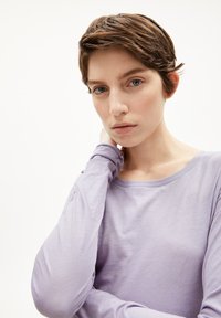Junge Person mit kurzen braunen Haaren, die ein lavendelfarbenes Langarmshirt trägt, hält sich mit einer Hand am Hals und schaut direkt in die Kamera.
