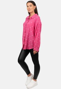 Camicetta rosa con pattern a cuori bianchi, abbinata a leggings neri e sneakers bianche. La camicia presenta una vestibilità comoda e maniche lunghe.