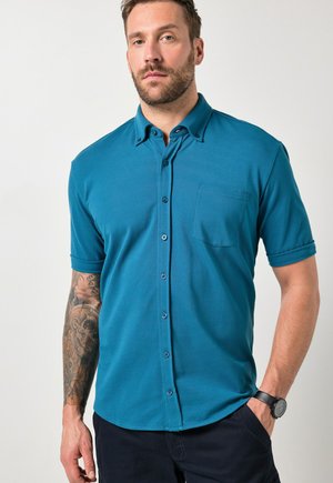 Mężczyzna ubrany w krótką koszulę na guziki w kolorze morskim, czarny zegarek na lewej ręce, z tatuażem na prawym przedramieniu, stoi na tle jednolitego białego tła.