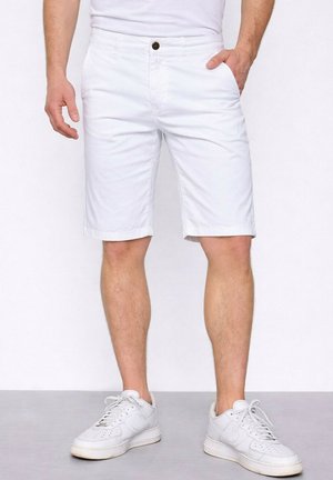 Homme portant un short blanc jusqu'aux genoux avec des poches avant et des baskets blanches, debout sur un sol gris contre un mur blanc.