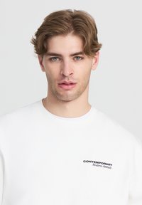 Felpa bianca con colletto rotondo e un piccolo logo ricamato nero sul petto sinistro con la scritta "CONTEMPORARY Modern Attitude."