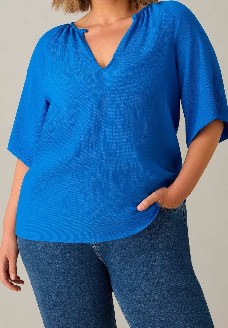 Frau trägt eine hellblaue, strukturierte Bluse mit V-Ausschnitt und Ellbogen-langen Ärmeln sowie dunkelblaue Jeans, die Hand in der Tasche ruhend.