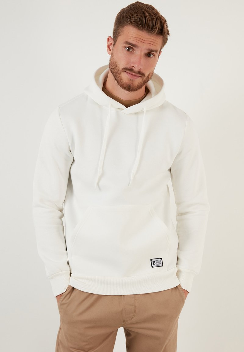 Bijela hoodie izrađena od mekog materijala, s prednjim džepom i povlačnim konopcima. Riblaste manžetne i donji rub, s malom markiranom etiketom sa strane.