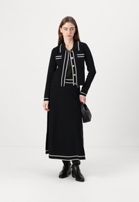 Black knit jacket with white lining and gold buttons, paired with a matching black skirt featuring white trim at the hem. Black boots and handbag.
