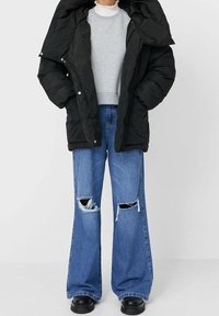 Sort polstret jakke med stort hætte, grå sweatshirt og brede blå jeans med slidte huller, parret med kraftige sorte støvler.
