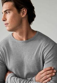 Grau geripptes Langarmshirt mit Rundhalsausschnitt. Der Stoff hat eine weiche Textur mit einem leichten Glanz. Die Arme sind verschränkt und teilweise sichtbar.