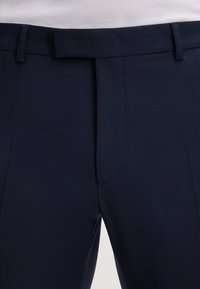 Marineblaue maßgeschneiderte Hose mit glatter Textur, flachem Vorderteil, scharfen Bügelfalten und Gürtelschlaufen. Der Stoff zeigt ein dezentes Webmuster.