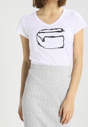 Vrouw draagt een wit T-shirt met V-hals met een zwart abstract ontwerp en een hooggetailleerde lichtgrijze geribbelde rok.