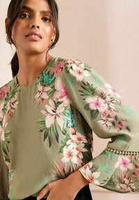 Floral blouse in olive green, featuring vibrant pink floral patterns, delicate butterfly accents, and textured bell sleeves with lace trim.