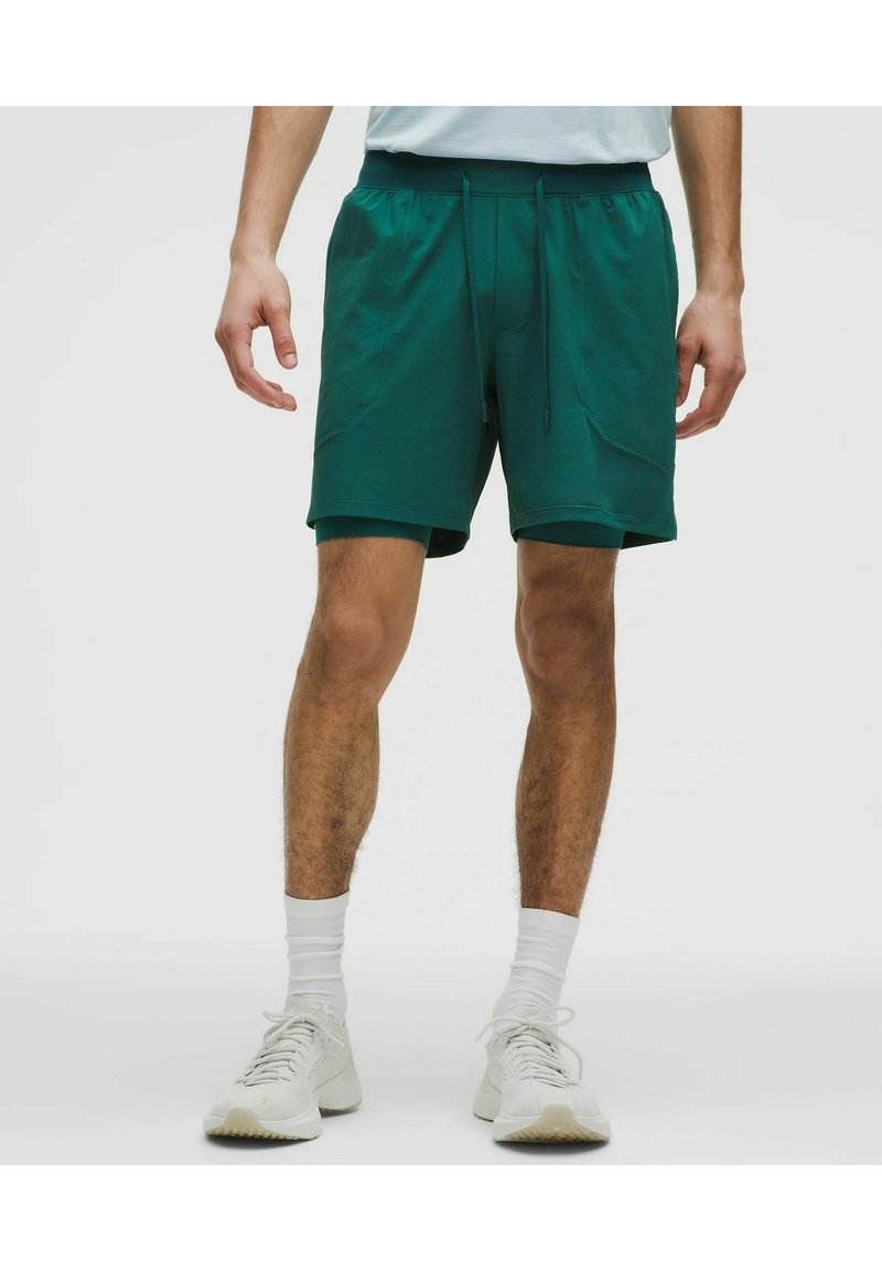 Man die donkergroene laagjessportshorts draagt, witte sokken en witte sneakers, staand met de armen ontspannen langs zijn lichaam.