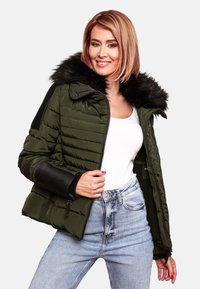 Giubbotto imbottito di colore verde scuro con colletto in pelliccia sintetica, dettagli a coste sulle maniche, tasche con zip e una vestibilità aderente su jeans chiari.