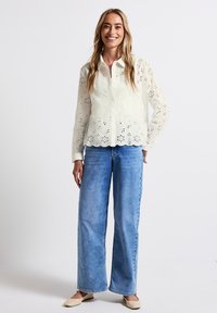 Vit broderad blus med scalloped fåll, i kombination med ljusblå vida jeans och beigefärgade lägenheter. Enkel krage och knäppning.
