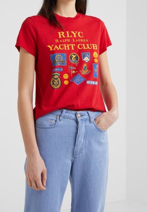 Kobieta ubrana w czerwoną koszulkę Ralph Lauren Yacht Club z naszywkami o tematyce żeglarskiej oraz jasnoniebieskie dżinsy, stojąca z jedną ręką w kieszeni.