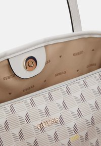 Biała torba na ramię z wzorem w geometryczny design, beżowe wnętrze z powtarzającym się logo "GUESS" oraz złote zapięcie na zatrzask.