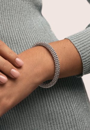 Brazalete de cuentas plateadas en una muñeca, con un diseño tejido y texturizado. La persona que lo lleva tiene las uñas de color rosa claro y viste una camiseta verde acanalada.