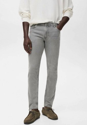 Man in een lichtgrijze slim-fit jeans, een witte langemouwen shirt en bruin suède veterschoenen, staande met één hand in zijn zak.