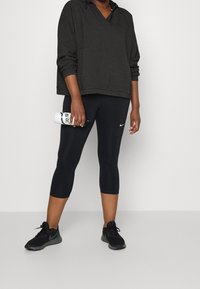 Zwarte gestructureerde hoodie gecombineerd met zwarte capri leggings. Houdt een witte waterfles vast met zwarte letters. Zwarte sportschoenen.