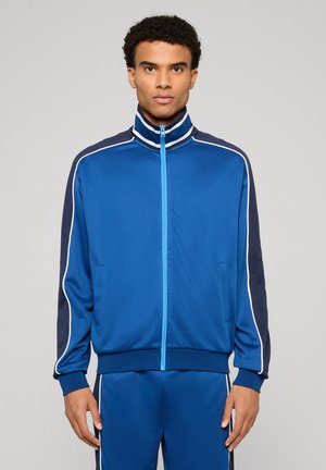 Mann trägt eine blaue Trainingsjacke mit Reißverschluss und passende Hose mit weißen und marineblauen Streifen, steht vor einem einfarbigen Hintergrund.
