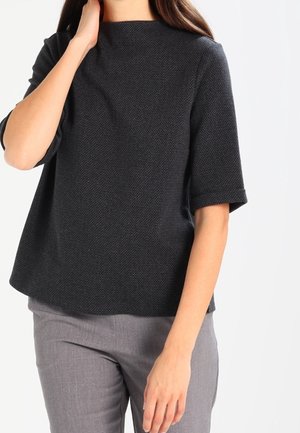Femme portant un haut texturé gris foncé à manches jusqu’aux coudes et un pantalon gris clair, se touchant le cou d’une main, de face.