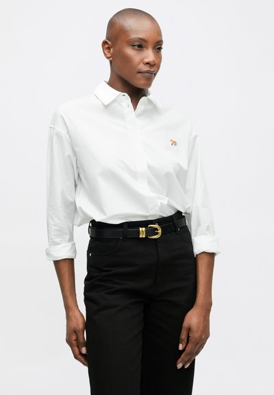 Biela košeľa na gombíky s vyhrnutými rukávmi a vyšívaným logom líšky, skombinovaná s čiernymi nohavicami s vysokým pásom a čiernym opaskom so zlatými doplnkami.