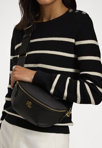 Schwarze Leder-Umhängetasche mit goldenen Beschlägen, Reißverschluss und geprägtem Logo. Getragen über einem schwarz-weiß gestreiften Pullover.