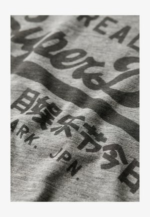 Tissu gris avec un texte et des graphismes noirs audacieux, présentant une apparence texturée et de légères plis, indiquant un design de T-shirt décontracté.