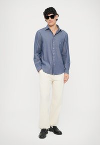 Camisa azul chambray abotonada con bolsillo, pantalones anchos beige claro, zapatos negros y gafas de sol. Textura suave, ajuste casual.