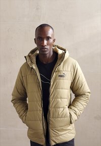 Giacca imbottita beige con colletto alto, chiusura zip e accento con logo. Indossata sopra una t-shirt nera, su uno sfondo neutro.