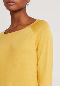 Pull tricot jaune avec un col large et des manches longues. Le tissu est lisse avec un léger éclat et présente des coutures raglan.