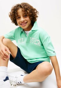 Polo shirt verde a maniche corte con testo del logo bianco, abbinato a pantaloni corti blu navy e scarpe da ginnastica bianche con accenti blu. Sfondo chiaro.