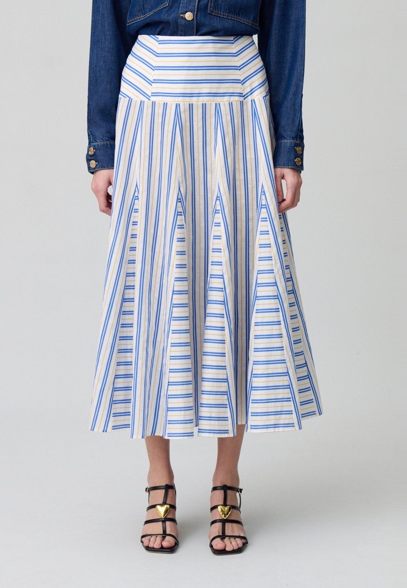 Falda midi a rayas en tonos crema, azul y dorado; cuenta con un dobladillo acampanado y pliegues verticales. Combinada con sandalias negras con detalles dorados.