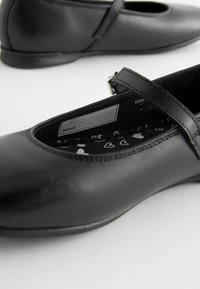 Svarta läderskor med platt sula, rund tå och rem över fotryggen; mjukt innerfoder med hjärtformade mönster; slät yta.