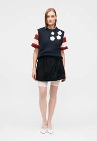 Marineblaues, ärmelloses Stricktop mit weißen Blumenapplikationen über gestreiften kurzen Ärmeln. Schwarzer Rock mit Spitzen-Unterhosen und Blumenakzenten. Weiße Schuhe mit Absatz.