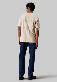 T-shirt in cotone beige con un grande logo CK ricamato sulla schiena, abbinato a jeans in denim blu scuro e scarpe nere.