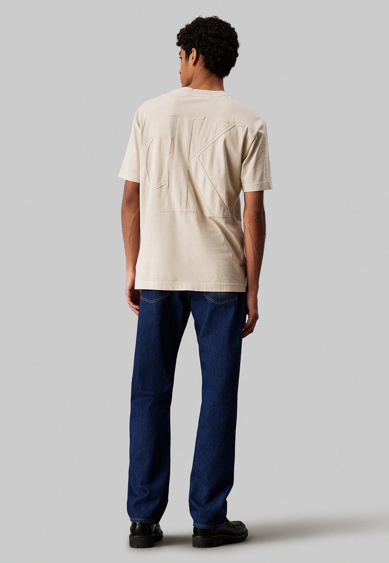 T-shirt en coton beige avec un grand logo CK brodé au dos, associé à un jean en denim bleu foncé et des chaussures noires.